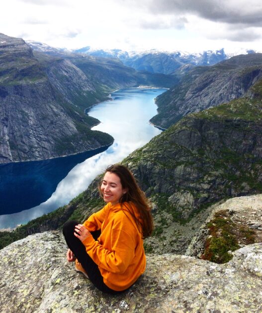 Woman sitting on the edge of the cliff, smiling, tourist hiking and exploring mountains in Norway